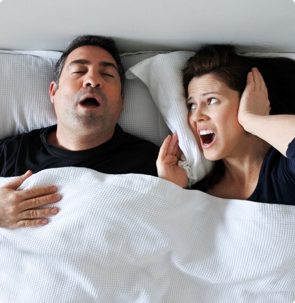 Frustrated woman covering ears in bed next to snoring man