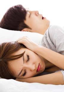 Frustrated woman covering ears with pillow while man snores next to her